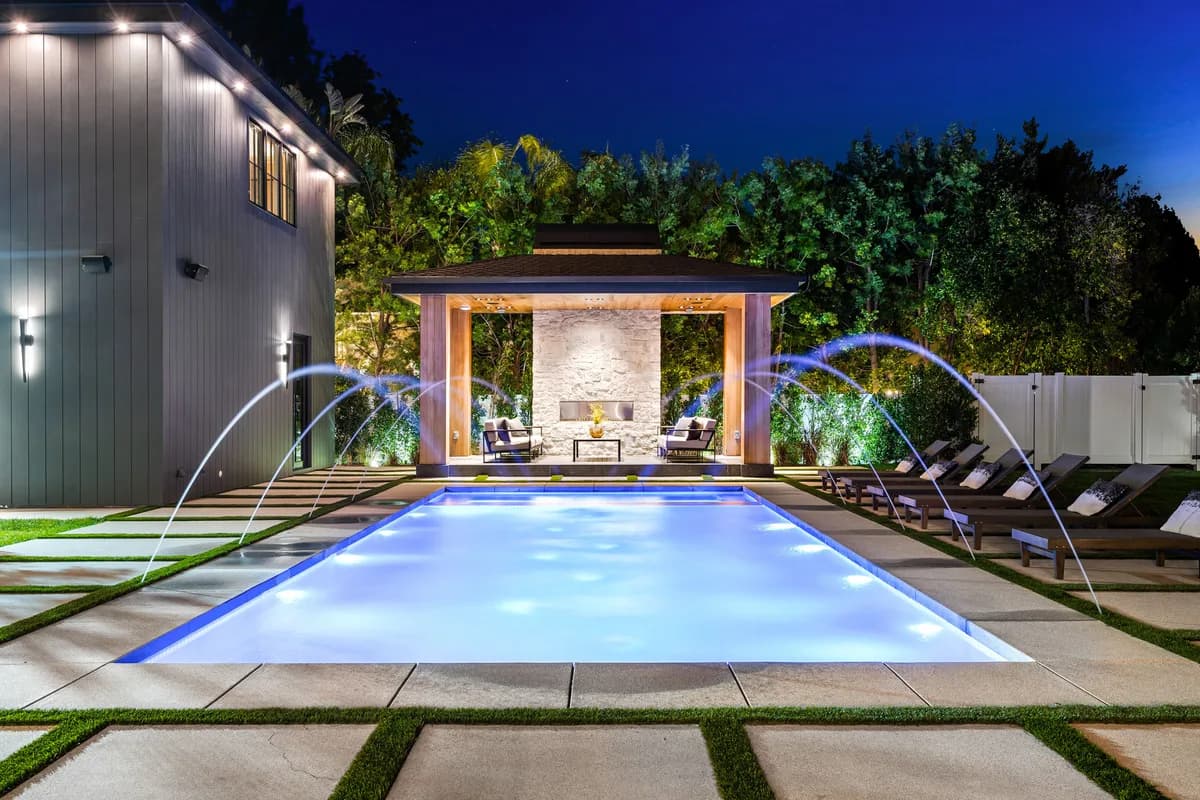Luxury residential pool illuminated at night with water jet arcs and a covered pavilion lounge area in the background
